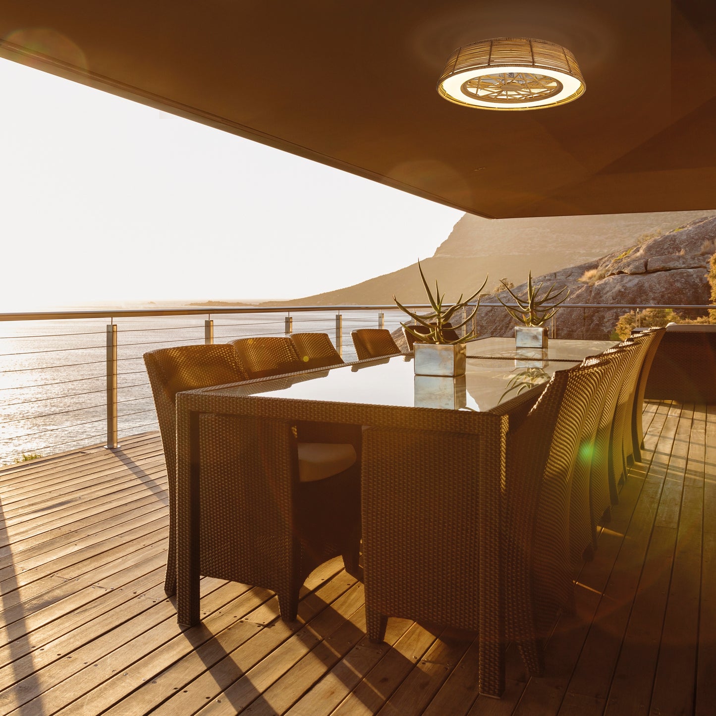 A sunlit patio with a wicker dining set and a long glass table overlooks the stunning ocean. Modern metal railings enhance the deck, while a Ceiling Fans UK Indonesia Mini Beige Ceiling Fan with Light adds charm. Small potted plants adorn the table, and mountains are visible in the distance.