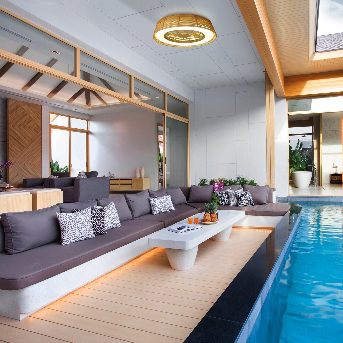 A contemporary indoor-outdoor living space showcases a sleek grey sectional sofa beside a pool. A white coffee table holds decorative items beneath an elegant Indonesia Mini Beige Ceiling Fan with Light, crafted by Ceiling Fans UK. This bright area features natural wood elements, expansive windows, and stylish design touches throughout.