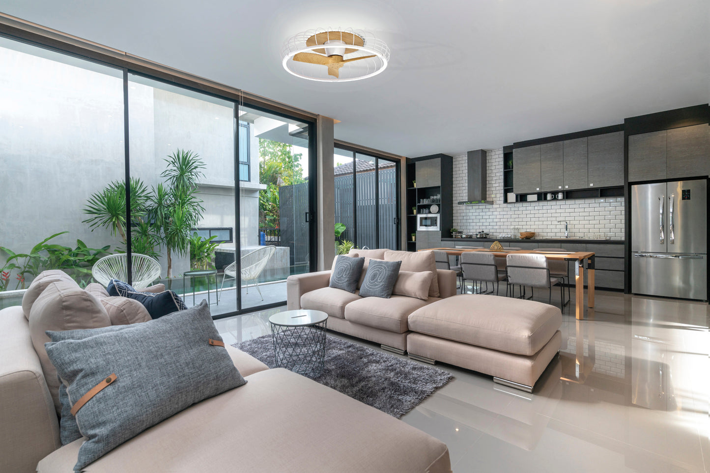 Modern open-plan living and kitchen space with large windows, beige sofas, and stainless steel appliances features a Ceiling Fans UK MANTRA M7809 Nature White/Beech 79cm LED dimmable ceiling fan with built-in DC reversible fan, remote & APP control.