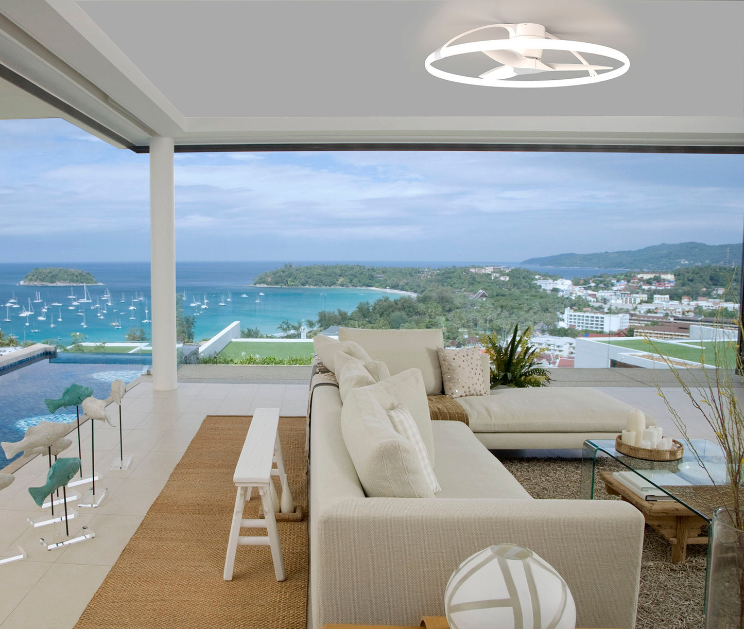 A modern, open living room features a light beige sectional sofa and a coffee table adorned with decorative items. A floor-to-ceiling window offers a view of the turquoise sea, boats, and a coastal town. The ceiling sports the stylish Nepal White Ceiling Fan with Light by Ceiling Fans UK, featuring a 105cm 75W LED dimmable light with a built-in 35W DC reversible fan, controlled via remote control, APP & Alexa/Google Voice Control, enhancing the contemporary ambiance.