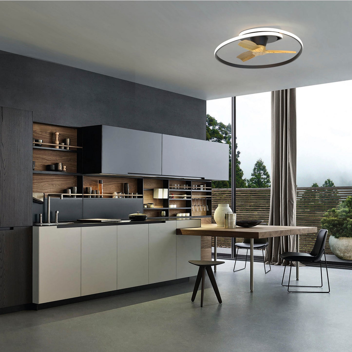 Modern kitchen with minimalist cabinets, open shelving, and sleek counters. A round dining table with mismatched chairs sits under the MANTRA M8236 Ocean Black/Wood Ceiling Fan with dimmable LED from Ceiling Fans UK. Large windows frame greenery in a neutral palette.