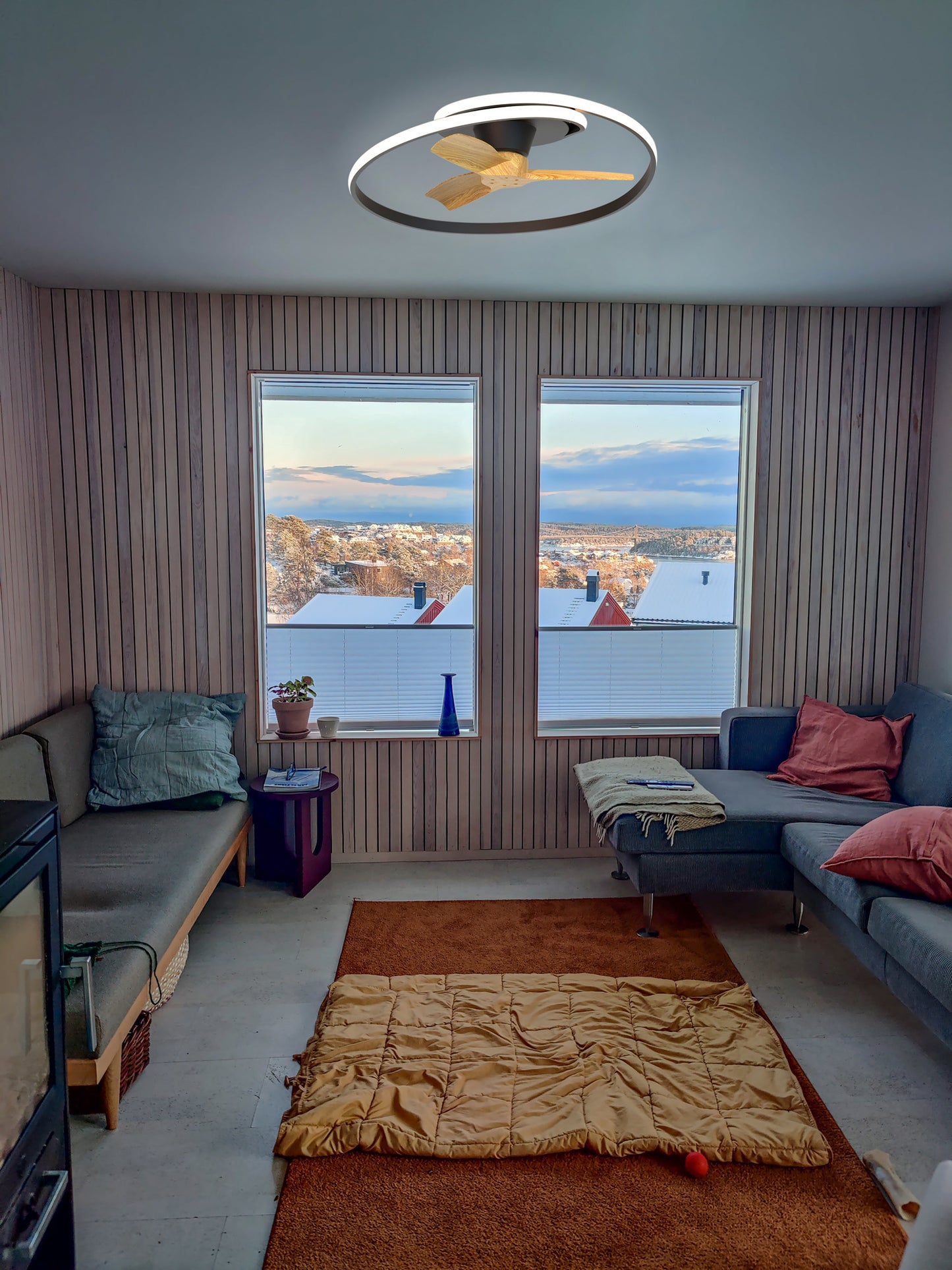 A cozy living room with wood walls, snowy views, gray sofas, colorful cushions, and a rug is brightened by the MANTRA M8236 Ocean Black/Wood Ceiling Fan with 60W dimmable LED light from Ceiling Fans UK—adding warmth and style.