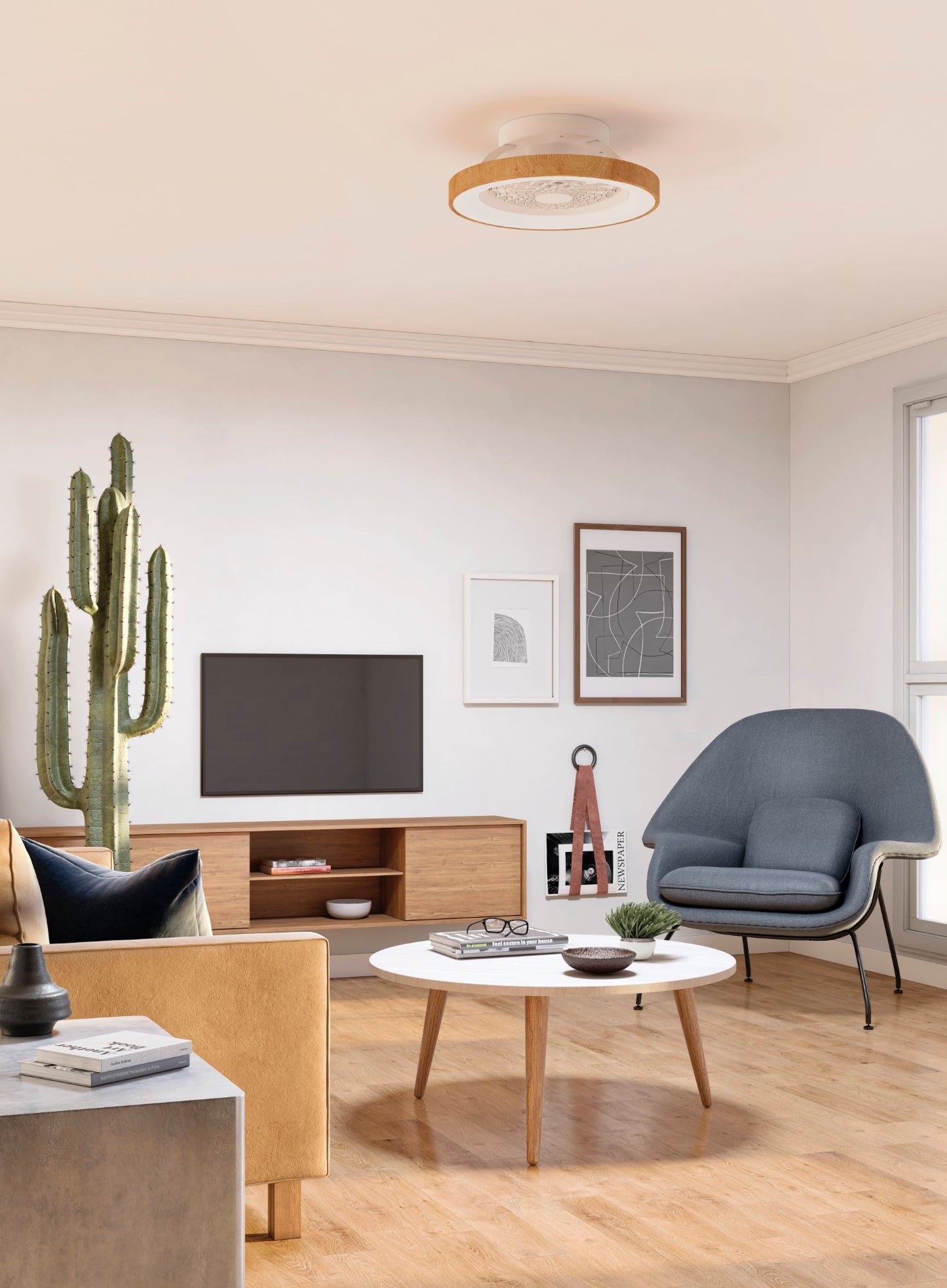 A modern living room boasts a wooden TV stand with a flat-screen TV, a blue armchair, a beige couch, and a round white coffee table. The Ceiling Fans UK Tibet Mini White/Wood Ceiling Fan with Light 53cm 70W LED Dimmable Ceiling Light With 35W DC Reversible Fan Remote controls the airflow overhead. A cactus plant, abstract wall art, and various decor items add character to the bright, minimalist space.