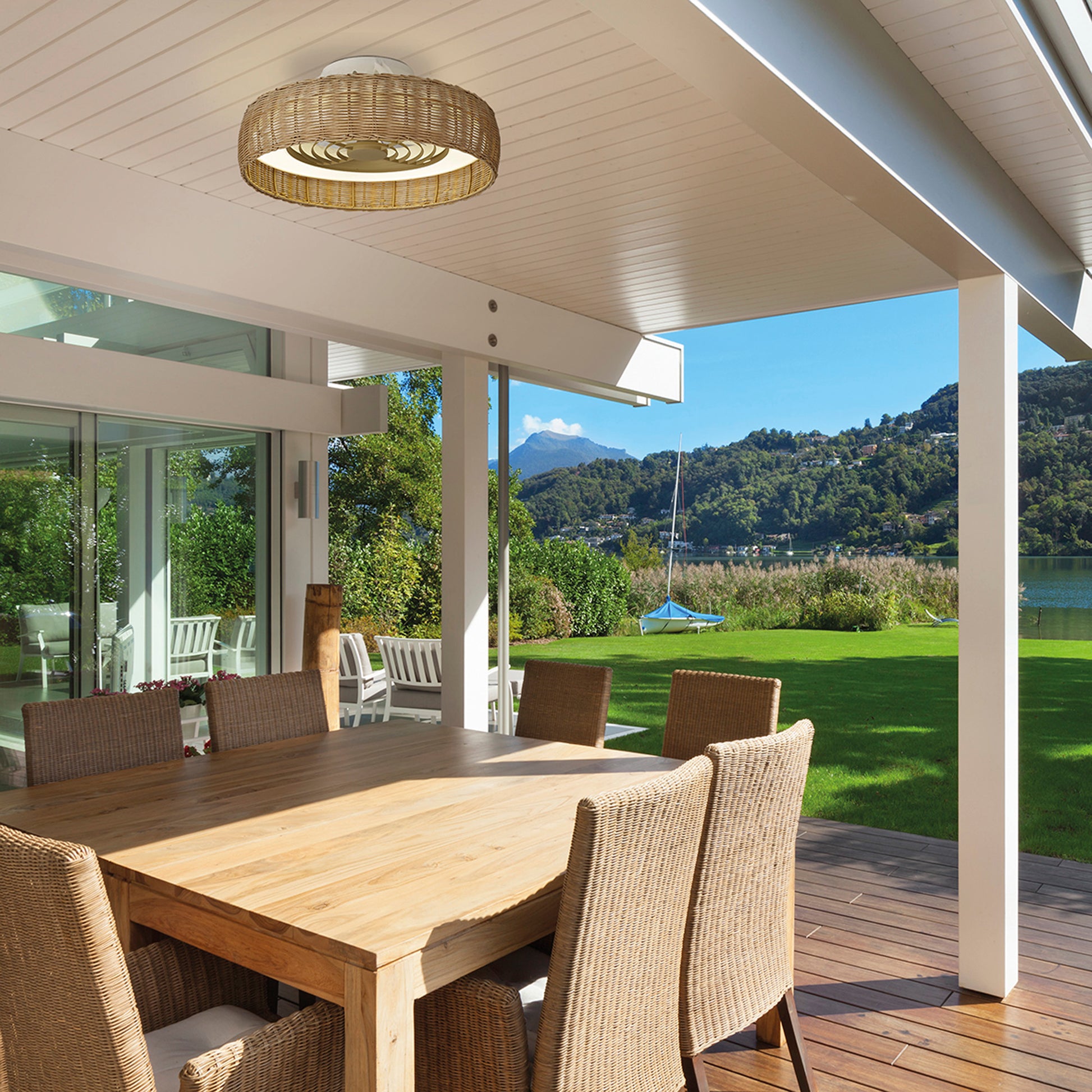 A covered patio features a wooden dining table and wicker chairs, lit by the Ceiling Fans UK Kilimanjaro Mini White Fan with Beige Rattan Shade 58cm 55W LED Dimmable Ceiling Light, overlooking a lawn, lake, and hills. Glass doors lead inside.