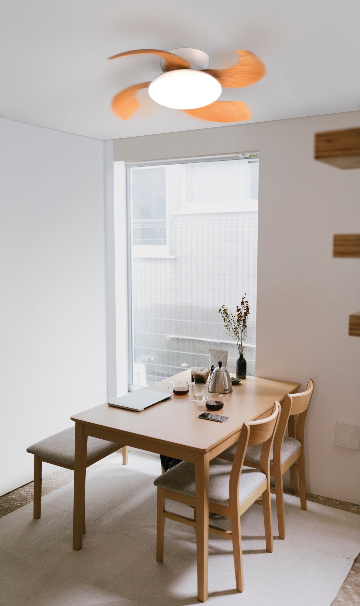 Minimalist dining area featuring a light wooden table with four matching chairs. A simple place setting includes a notebook, an open book, a teapot, and two cups. Above, the Ceiling Fans UK Aloha White/Wood Ceiling Fan with Light 52cm 45W LED Dimmable Ceiling Light With Built-In 30W DC Reversible Fan provides illumination and air circulation. Natural light streams through a large window.