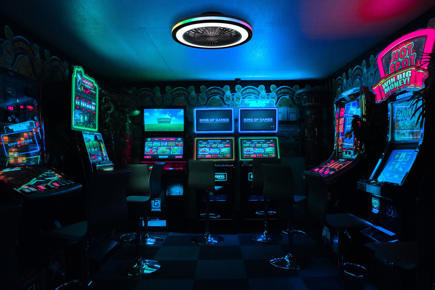 A dimly lit arcade with vibrant neon lights illuminates various slot machines and video game screens. The room has a futuristic ambiance with modern chairs and a Ceiling Fans UK Gamer Small Black Ceiling Fan featuring 40W LED dimmable white/RGB lighting and a built-in 20W DC reversible fan, complete with remote control. The walls are adorned with graffiti-style art, enhancing the edgy atmosphere.