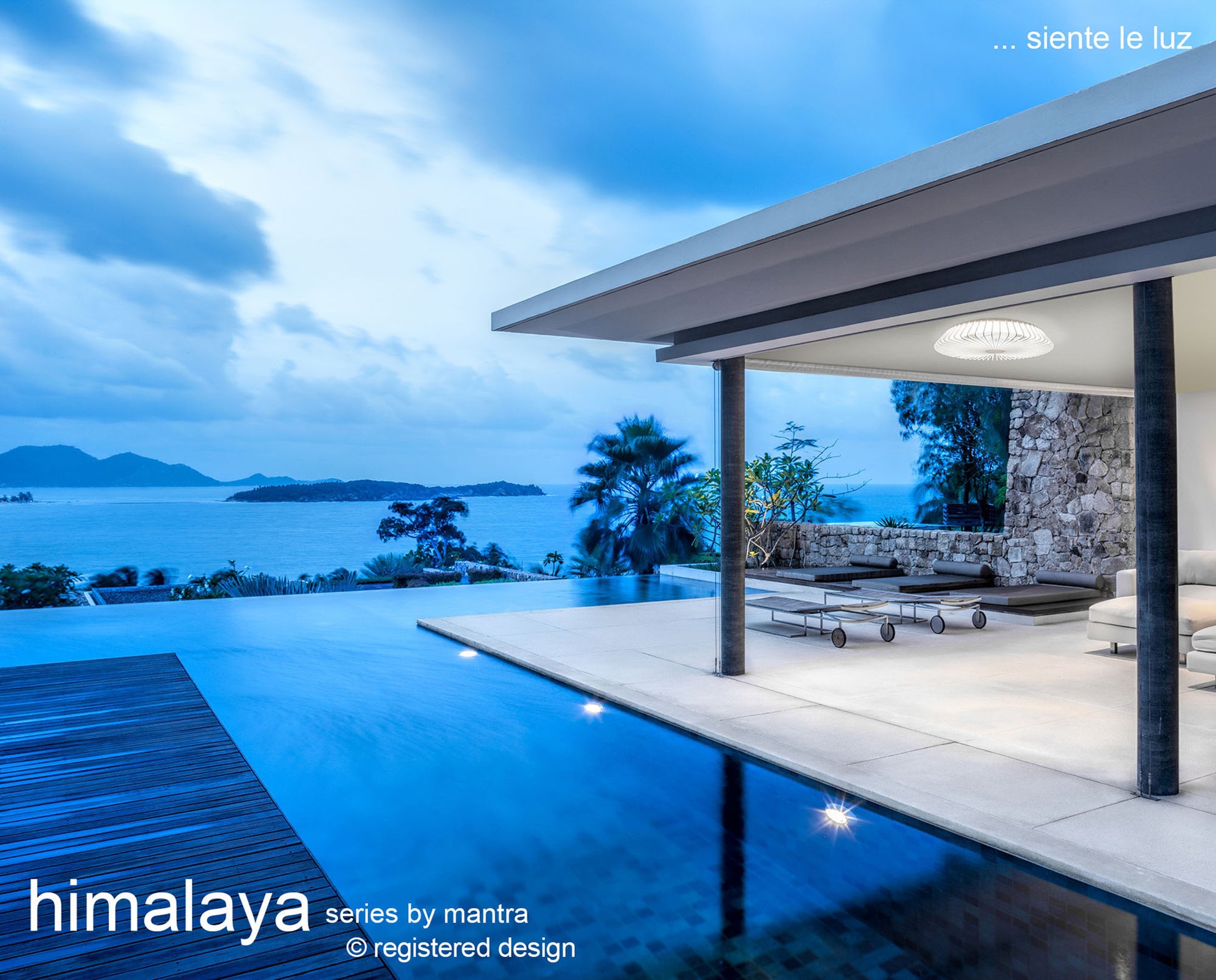 A modern waterfront patio provides a tranquil view of the calm blue sea and distant islands, featuring an enticing pool and cozy seating. The space is beautifully enhanced by the Himalaya White Ceiling Fan with Light from Ceiling Fans UK, standing out with its 63cm 70W LED Dimmable Light, reversible fan, remote control options including APP & Alexa/Google Voice Control, and an impressive 5-year warranty.