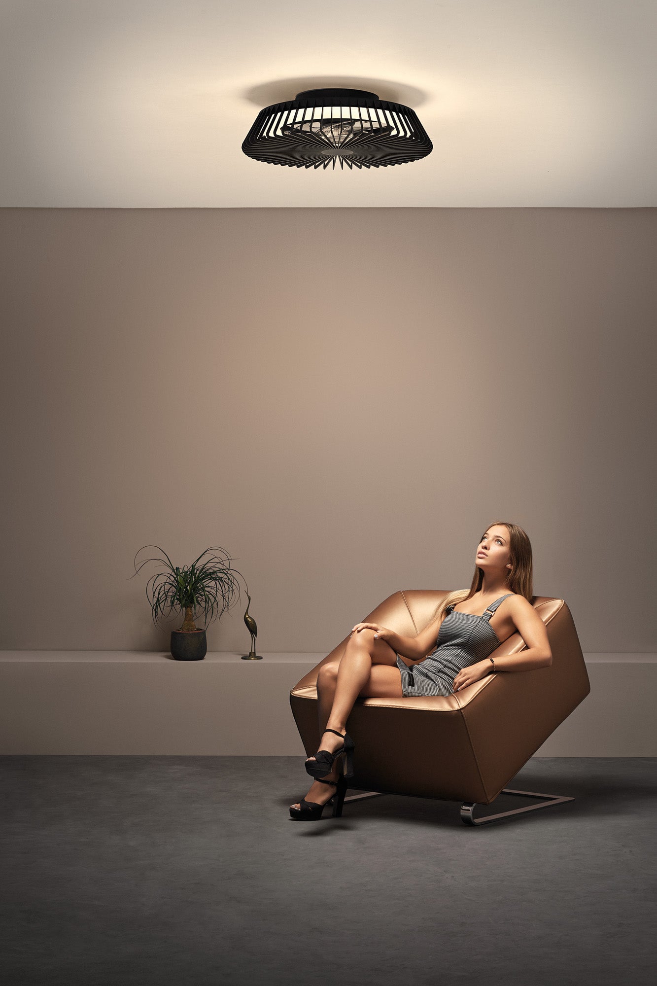 A woman in a gray dress sits in a modern, angular tan armchair. She looks upward at the Ceiling Fans UK Himalaya Mini Black Ceiling Fan with Light 53cm, featuring a 70W LED round ceiling light and a 30W DC reversible fan with remote control. A small potted plant and a decorative bird figurine are placed on a shelf in the background against a beige wall. The setting is minimalistic.