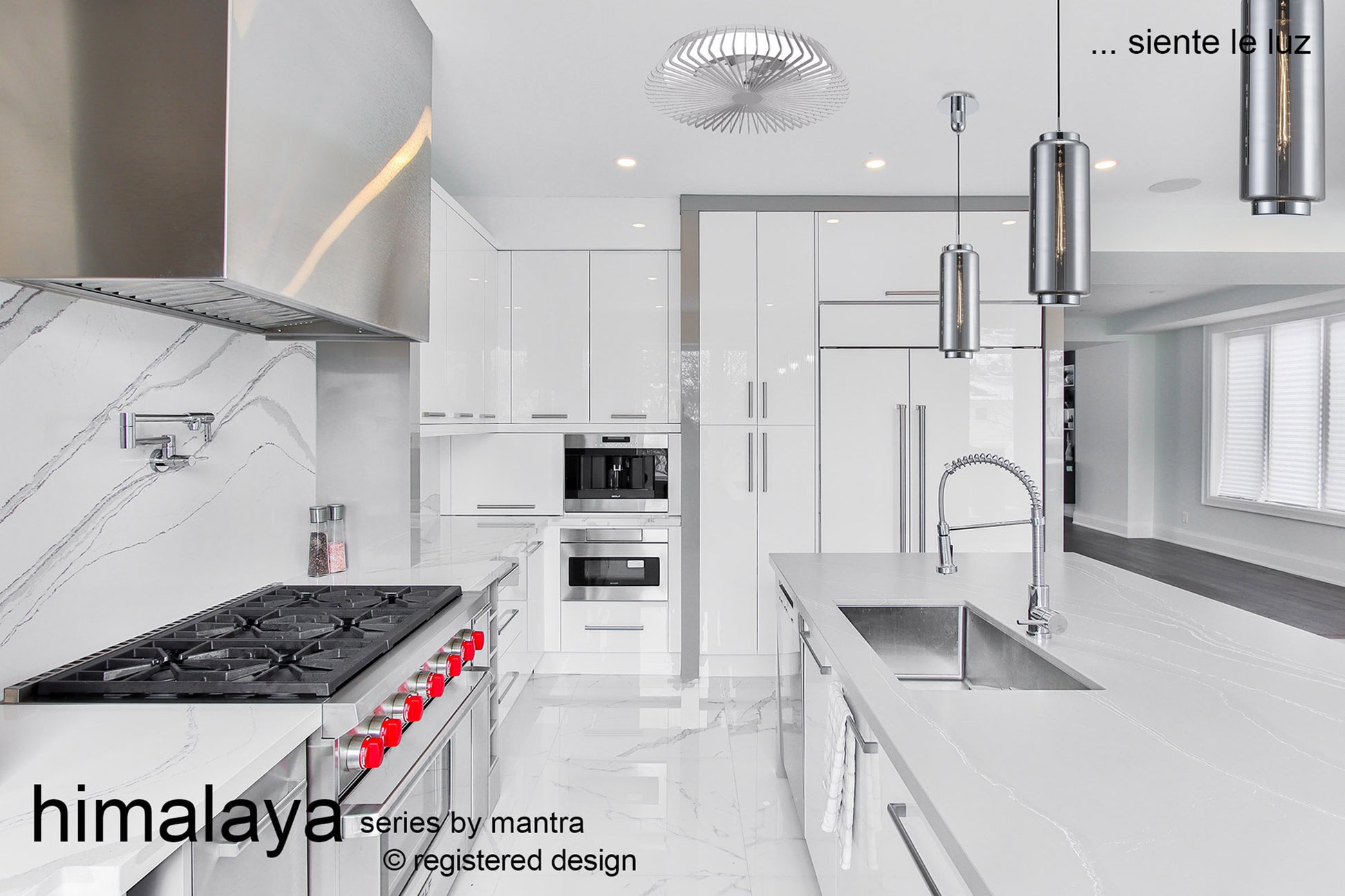 A modern, minimalist kitchen in white with stainless steel appliances features a marble island countertop with a built-in sink and faucet, gas stove with red knobs, an LED round ceiling light from Ceiling Fans UK, pendant lighting, and a sleek design. Text in Spanish includes "Himalaya Mini White Ceiling Fan" and "siente la luz," among other details.