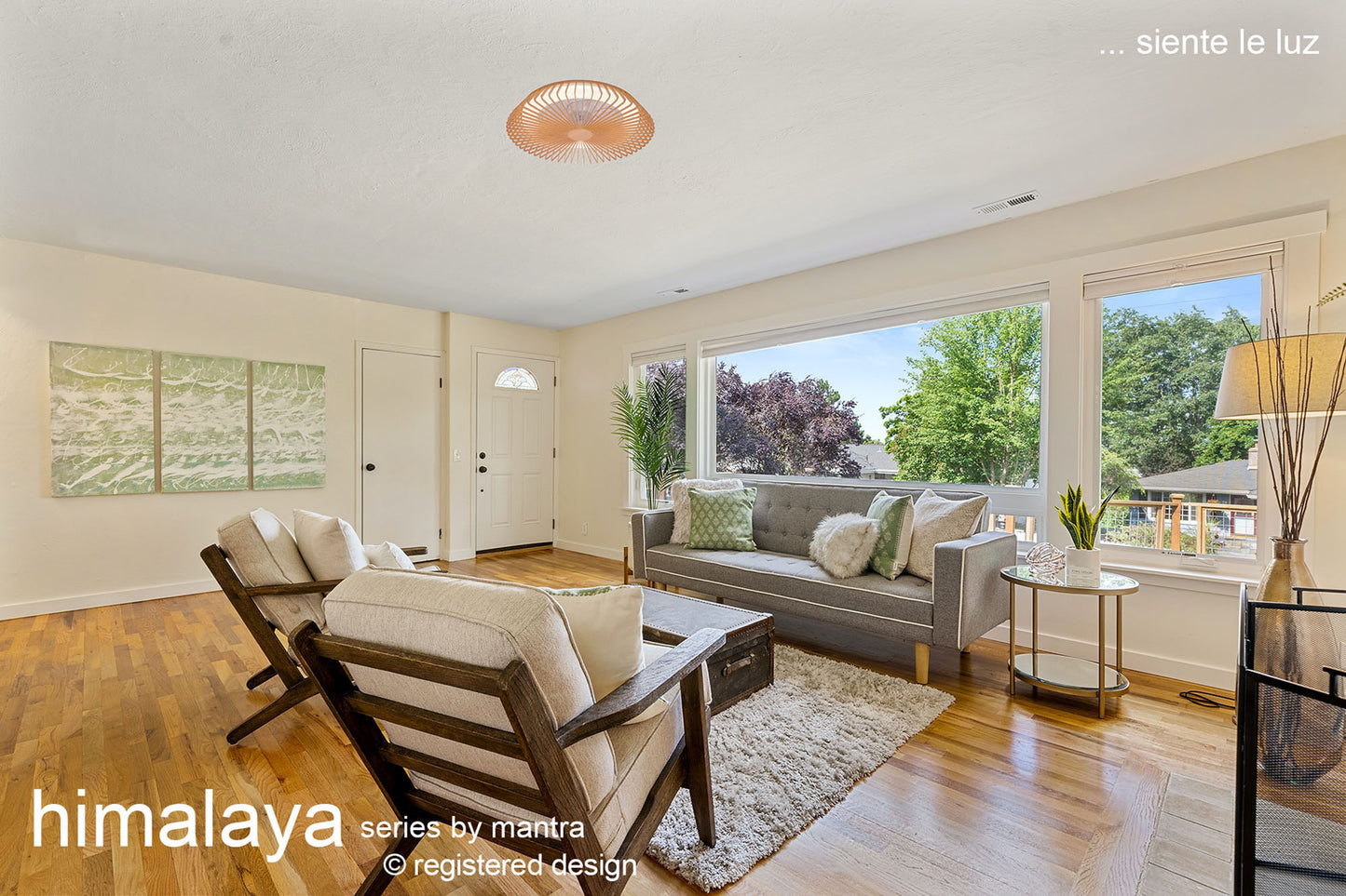 A modern, bright living room with a large window lets in natural light. It features wooden floors, two armchairs, a gray sofa, and a small round table. A wall art piece is mounted on the left, while the Ceiling Fans UK Himalaya Wood Ceiling Fan with Light 63cm 70W LED Dimmable adds elegance. Lush greenery is visible outside.