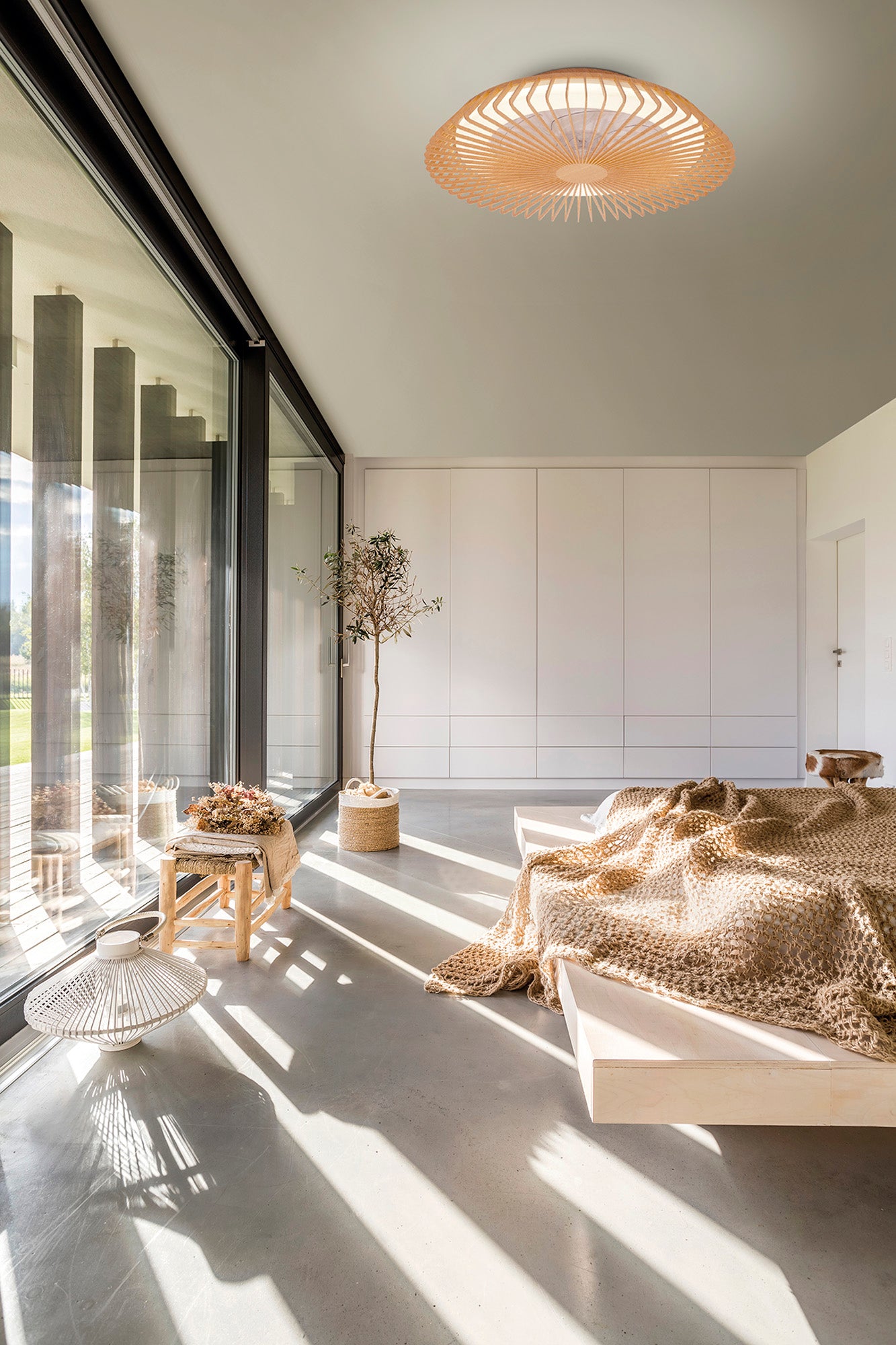 A minimalist bedroom with expansive floor-to-ceiling windows showcases a low wooden bed adorned with a textured throw, accompanied by a potted plant and a wicker chair with cushions. The Himalaya Wood Ceiling Fan with Light from Ceiling Fans UK offers LED dimmable lighting, while sunlight streaming in casts captivating shadows on the floor.