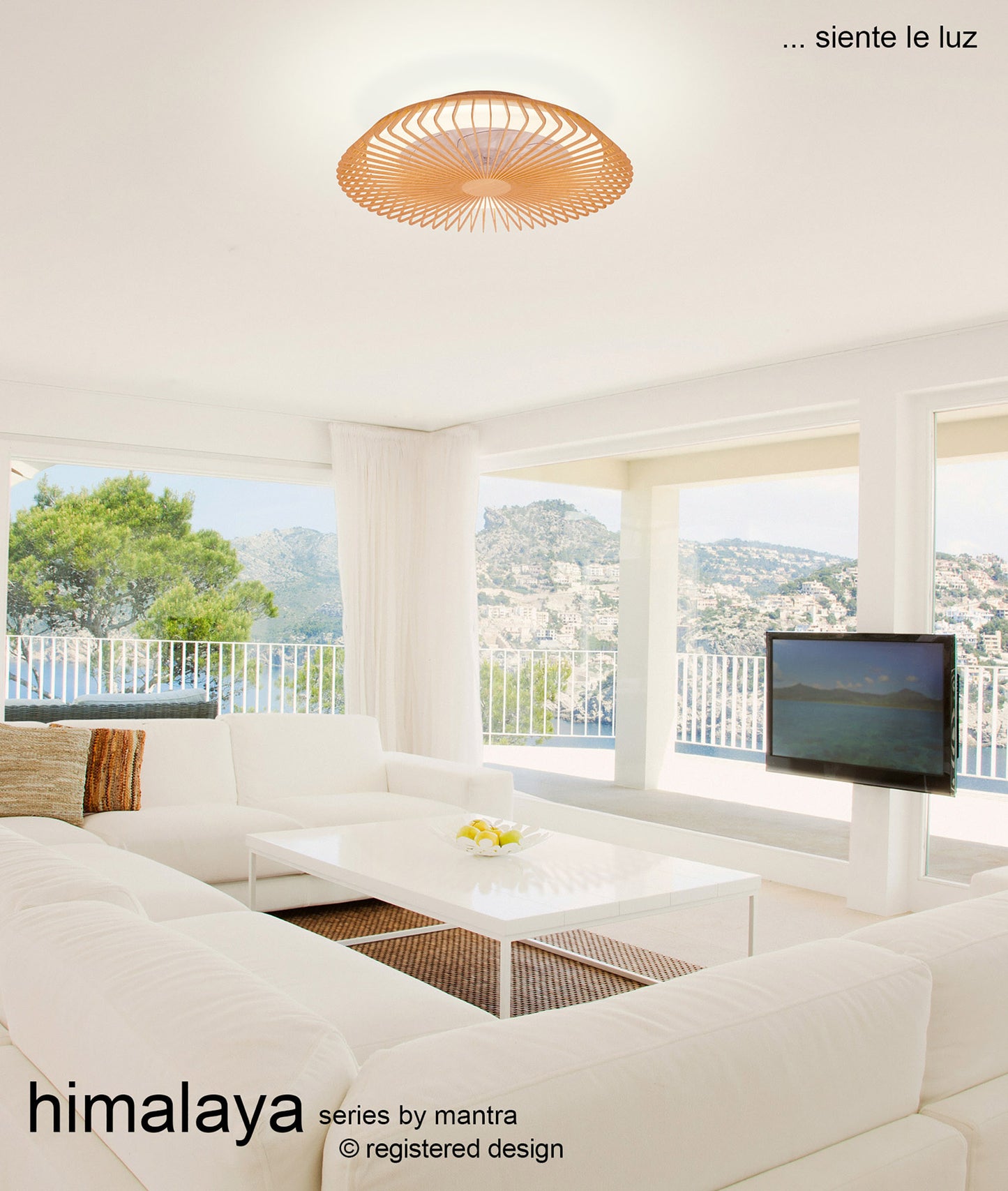 A modern, bright living room featuring a white sectional sofa and a white coffee table. The Himalaya Wood Ceiling Fan with Light (63cm 70W LED Dimmable Light With Built-In 35W DC Reversible Fan) by Ceiling Fans UK adds elegance to the decor, while large windows offer a scenic view of a hillside landscape. Text reads "Himalaya Series by Mantra" and "... siente le luz.
