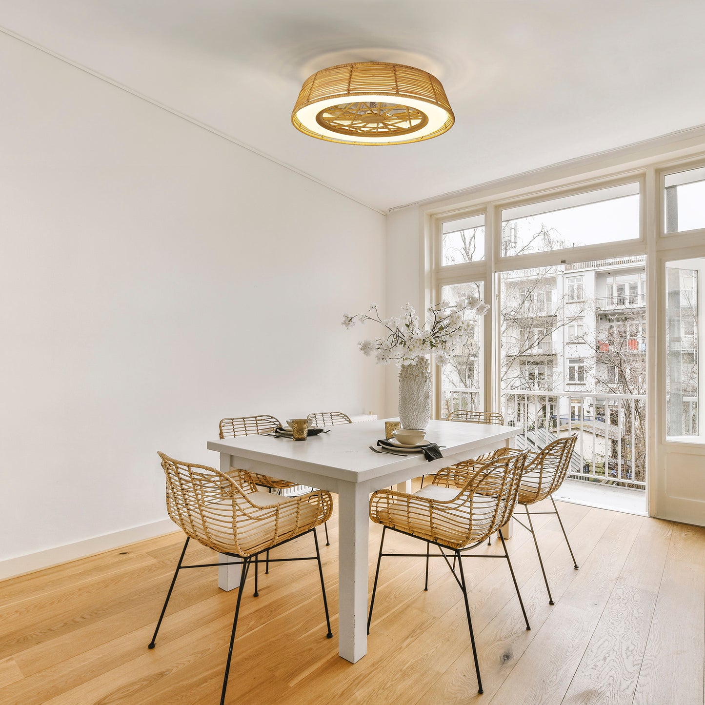 A bright dining room with a minimalist design showcases a white table encircled by six woven chairs. The Indonesia Beige Rattan Ceiling Fan with Light by Ceiling Fans UK complements the basket-style light fixture above, its beige rattan finish enhancing the decor. A large window bathes the space in natural light, accentuating the hardwood floor.