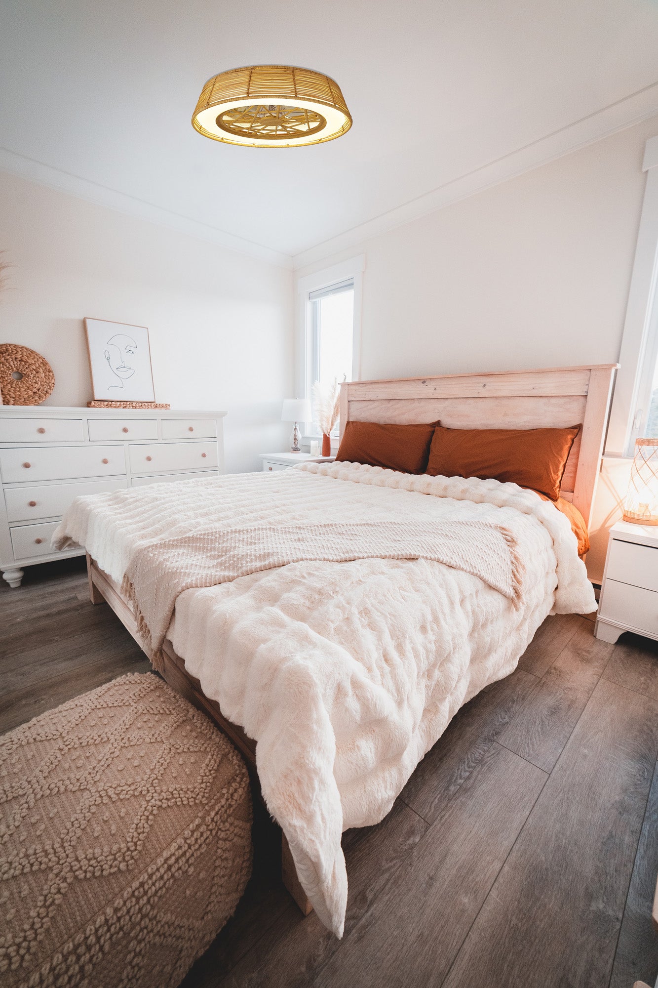 A cozy bedroom features a wooden bed with a fluffy beige comforter and orange pillows. A white dresser holds a framed art piece, while the room's wood flooring is softly illuminated by the "Indonesia Beige Rattan Ceiling Fan with Light" from Ceiling Fans UK, complete with a 70W LED dimmable light function. A small pouf sits at the foot of the bed.
