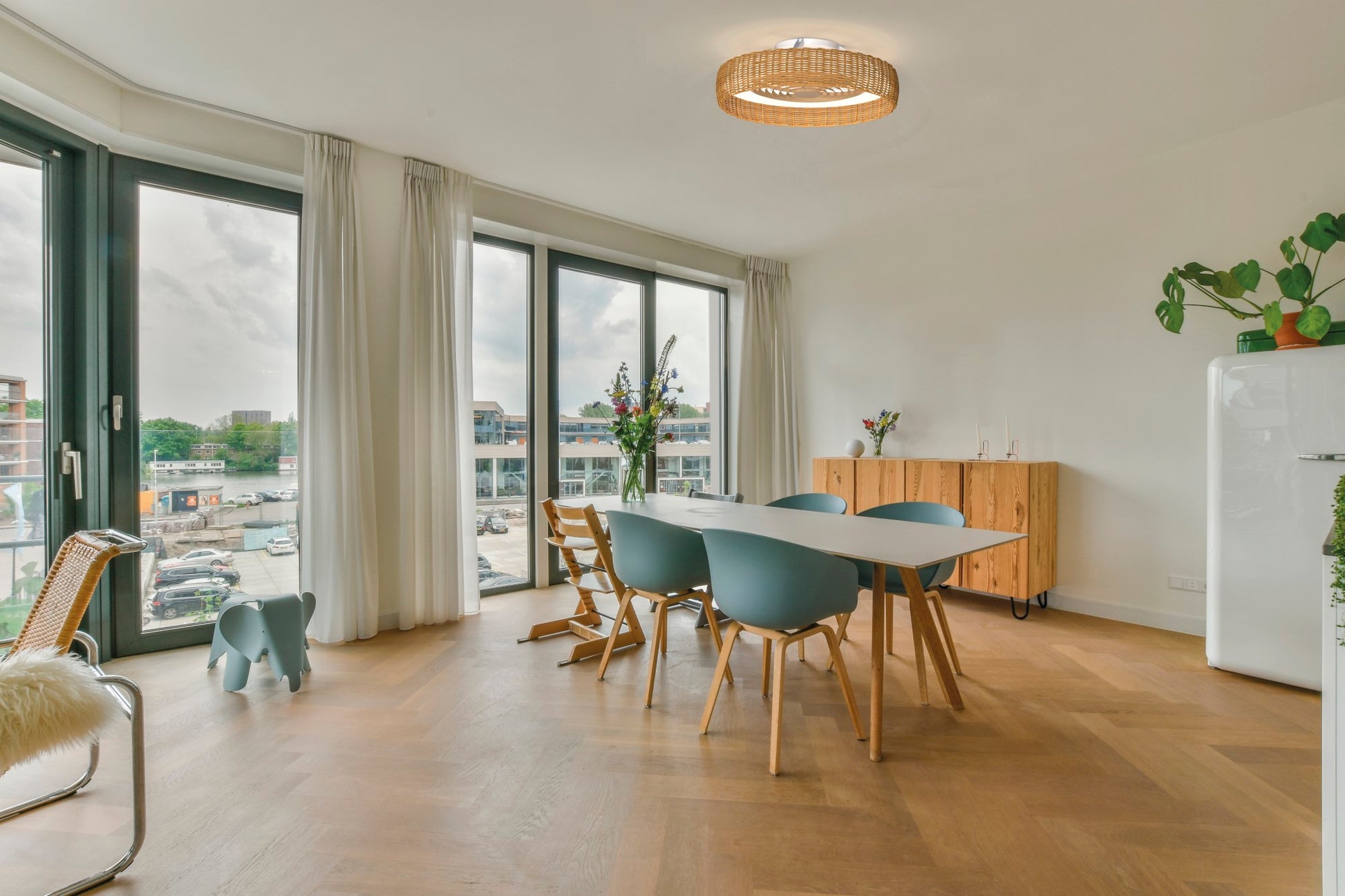 A modern dining room with large windows allows natural light to fill the space. It features a wooden table with blue chairs, a wooden sideboard, and potted plants. The Kilimanjaro White with Beige Rattan Shade ceiling fan by Ceiling Fans UK provides dimmable LED lighting to highlight a wicker chair with a fur throw and a charming blue elephant-shaped stool.
