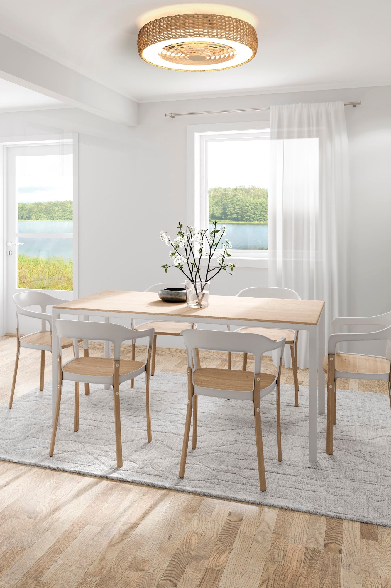 A bright dining room features a wooden table with white modern chairs on a light gray rug. A vase with blooming branches sits on the table. Large windows reveal a scenic view of a lake and trees. Hanging from the ceiling is the Kilimanjaro White Fan with Beige Rattan Shade by Ceiling Fans UK, offering dimmable LED lighting and remote-controlled reversible fan functionality. Sheer white curtains adorn the windows.