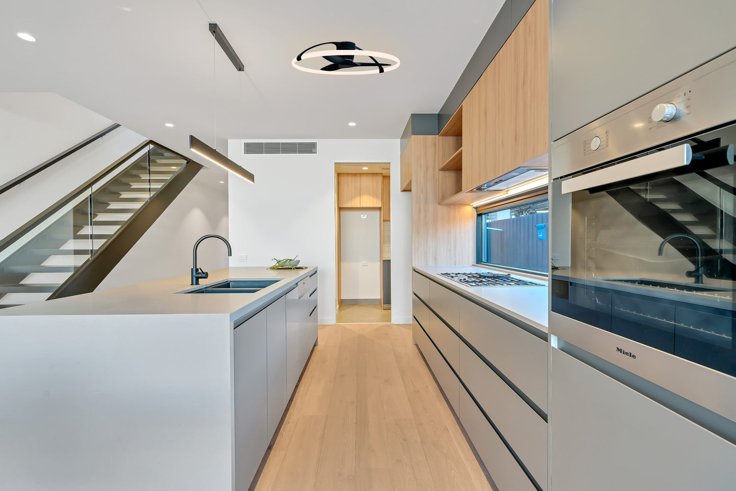 Modern kitchen features light wood floors, gray cabinetry, an island sink, built-in oven, and under-cabinet lighting. The MANTRA M7532 Nepal White/Beech Ceiling Fan with 75W dimmable LED by Ceiling Fans UK adds ambiance and smart control. Laundry visible in back doorway.