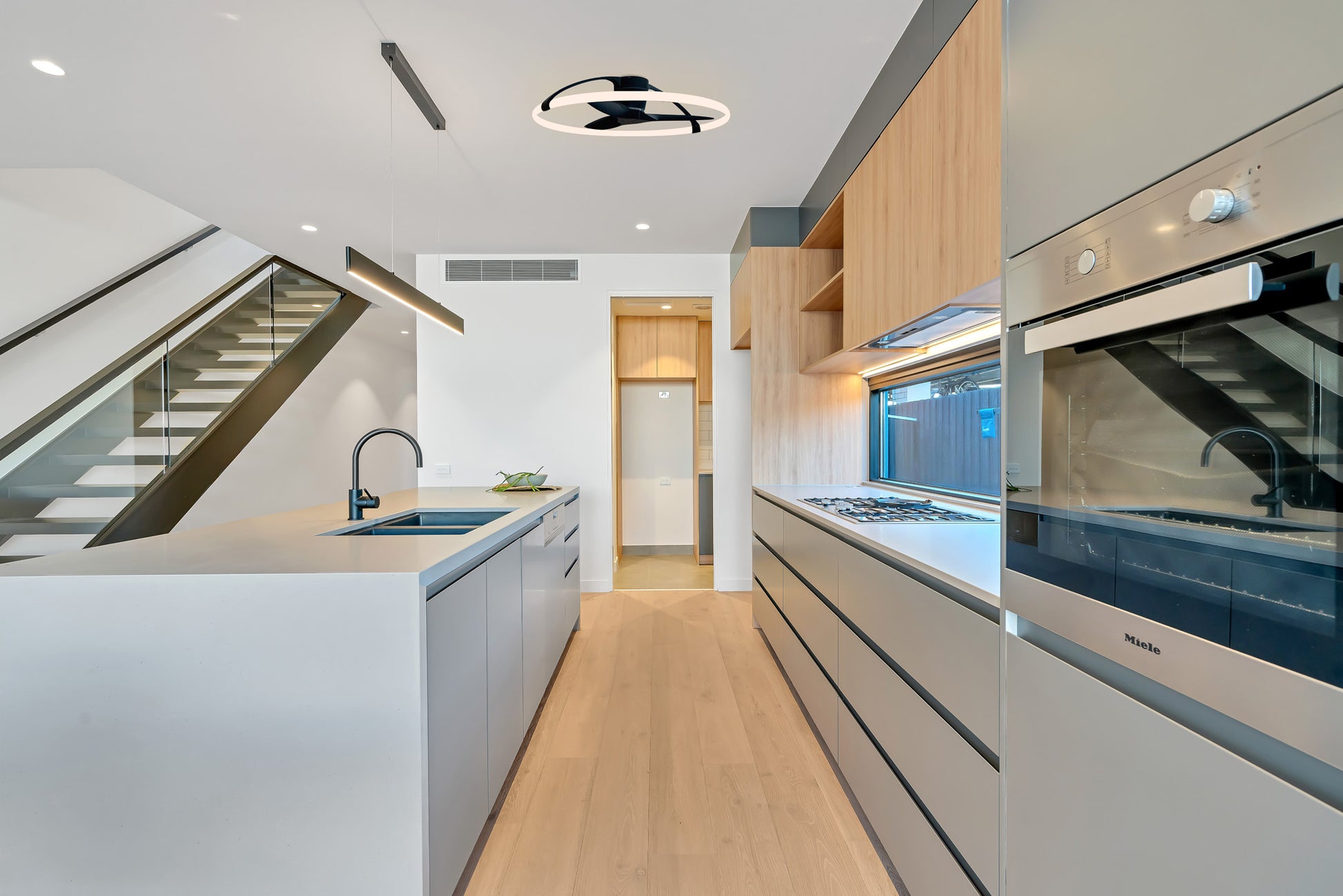 Modern kitchen features light wood floors, gray cabinetry, an island sink, built-in oven, and under-cabinet lighting. The MANTRA M7532 Nepal White/Beech Ceiling Fan with 75W dimmable LED by Ceiling Fans UK adds ambiance and smart control. Laundry visible in back doorway.
