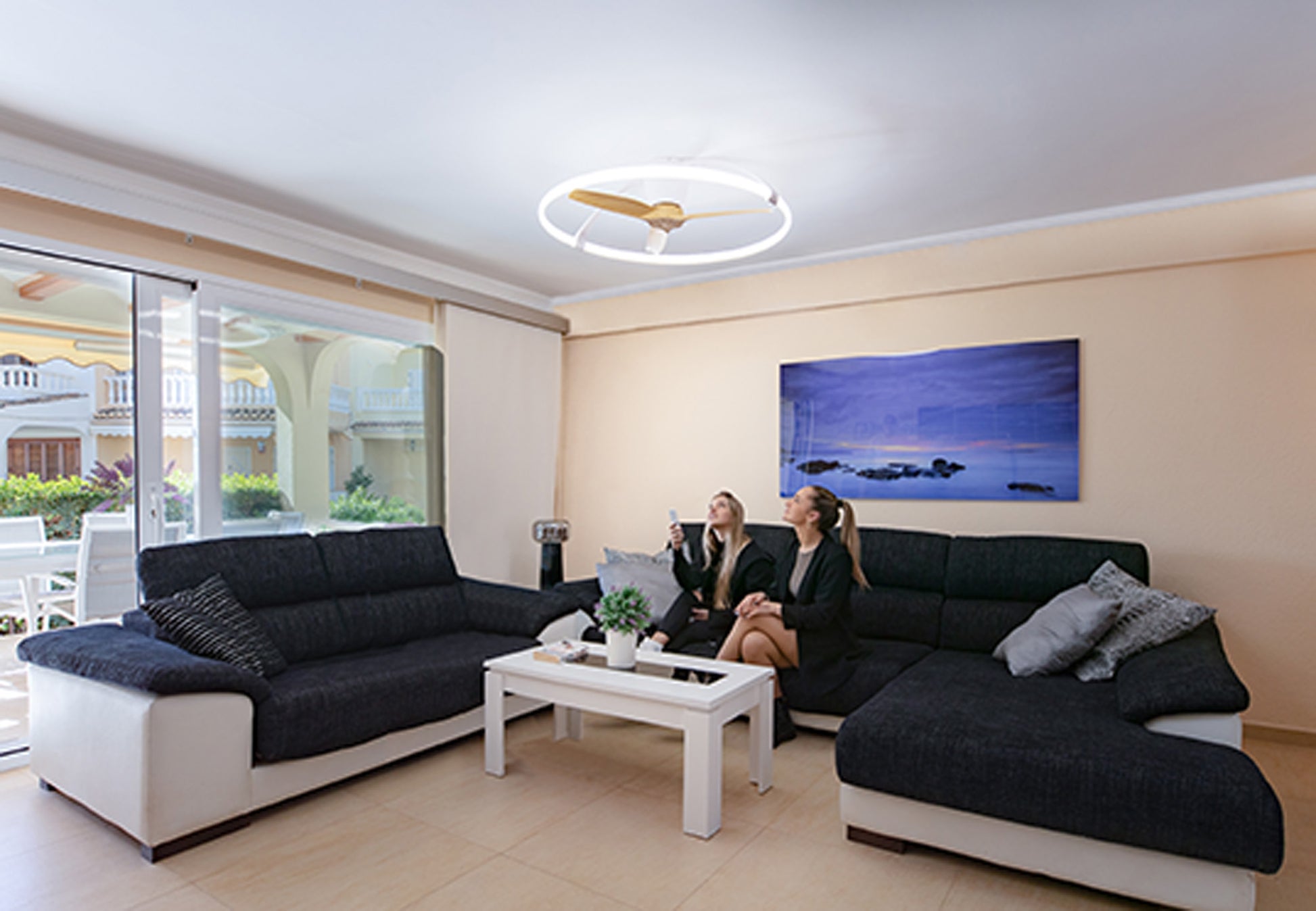 A modern living room featuring two women sitting on dark gray sofas with white accents, chatting. A white coffee table with plants and decor is placed between the sofas. The room has a large window with sheer curtains, a mounted blue wall painting, and a Nepal White/Beech Ceiling Fan with light 105cm 75W LED Dimmable Light With Built-In 35W DC Reversible Fan by Ceiling Fans UK overhead.