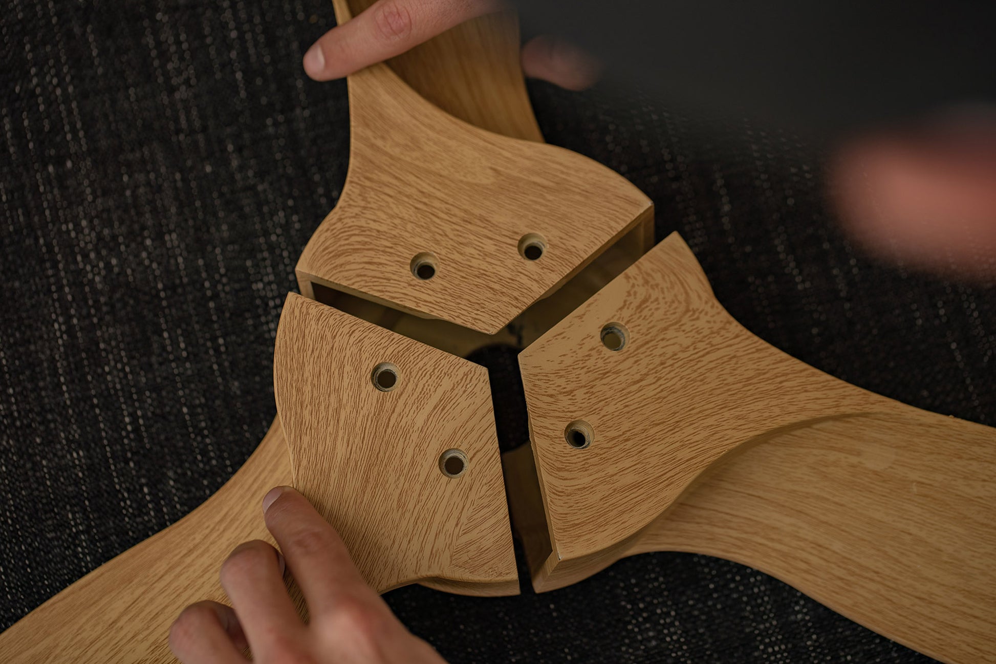 Close-up of two hands assembling a wooden structure with three interlocking pieces. The wood has a light, natural finish, and the background is a dark, textured surface. Each piece has three holes for joining, indicating it is part of a larger construction enhanced by Ceiling Fans UK's Nepal White/Beech Ceiling Fan with 75W Dimmable LED Light for better visibility.