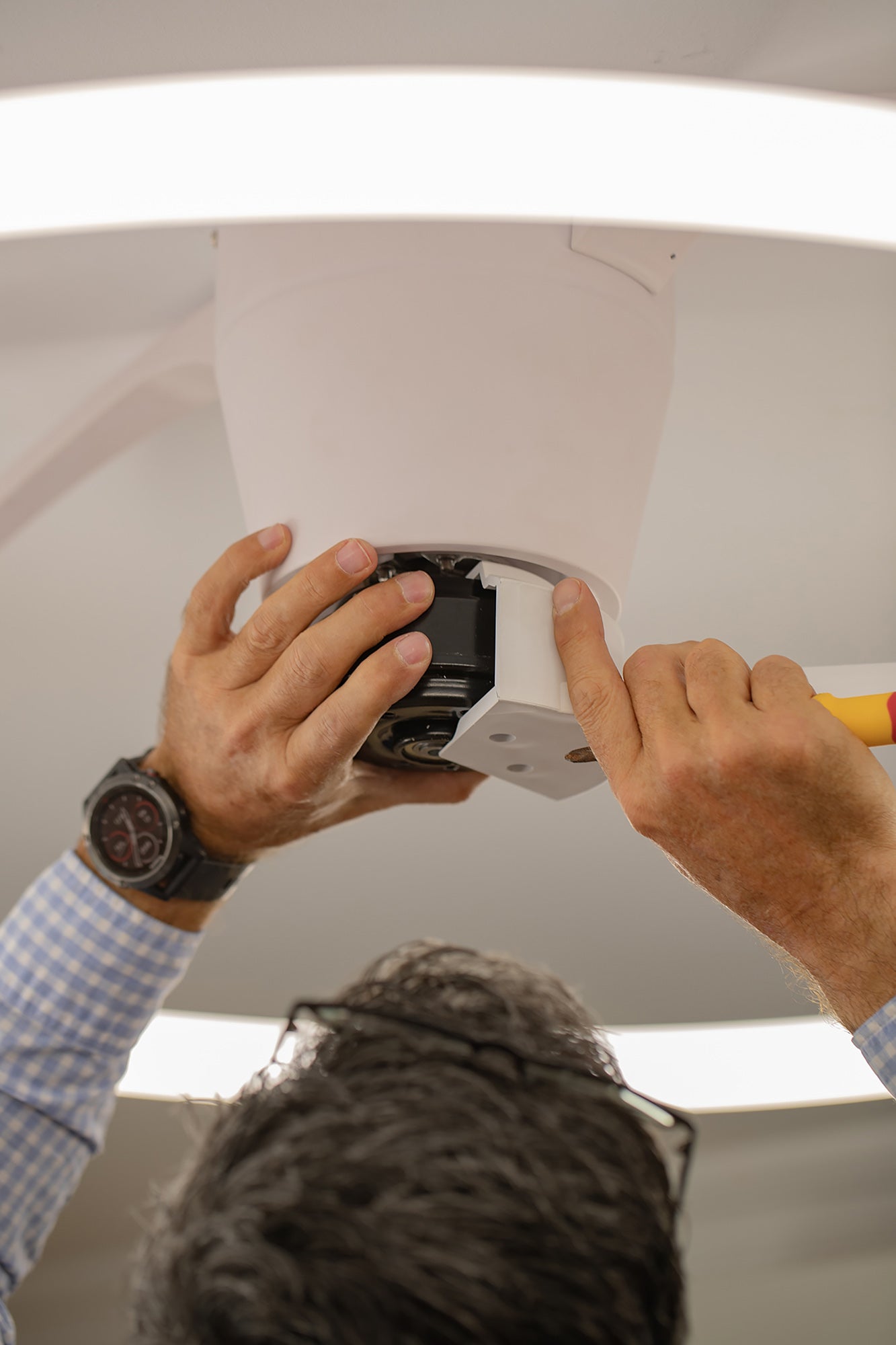 A person wearing a blue checkered shirt and a wristwatch is installing or repairing a Nepal White/Beech Ceiling Fan with LED light on the ceiling. Their hands are adjusting the device, and part of the person's head and glasses are visible from behind.