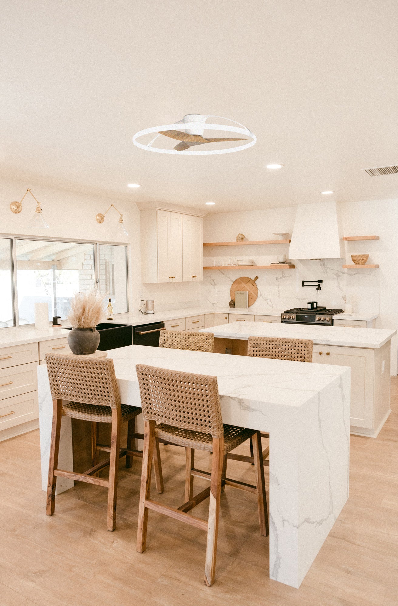 A modern kitchen features a marble island with two wicker bar stools, light wood flooring, white cabinetry, and stainless steel appliances. A decorative arrangement sits on the island, and the space is well-lit by large windows and the contemporary Ceiling Fans UK Nepal White/Beech Ceiling Fan with light 105cm 75W LED Dimmable Light With Built-In 35W DC Reversible Fan, Finish c/w Remote Control, APP & Alexa/Google Voice Control, offering an impressive 7200lm.