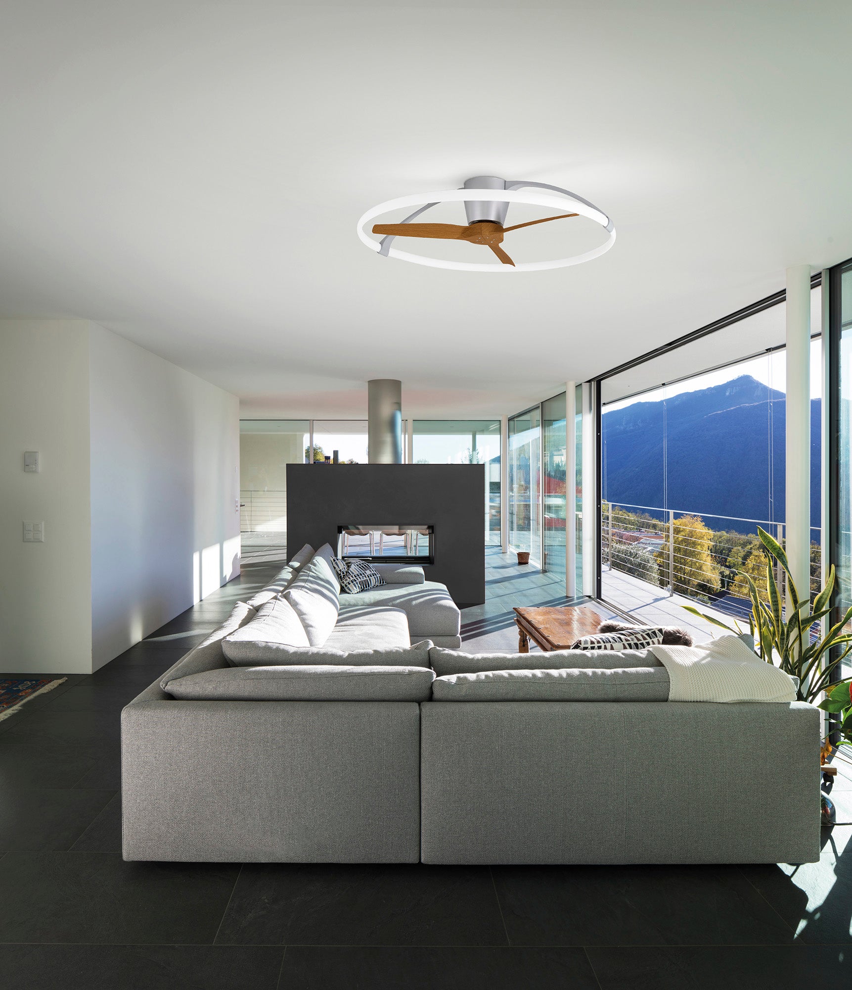 A modern living room features a large gray sectional, mountain views through glass walls, and a wooden coffee table. The Ceiling Fans UK MANTRA M7533 Nepal Silver/Walnut ceiling fan offers dimmable 75W LED light and smart voice/app control.