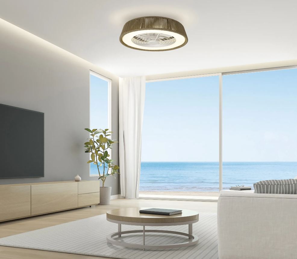 A modern living room features a round coffee table, a white couch, and a potted plant. Expansive floor-to-ceiling windows showcase stunning ocean views. Mounted above is the Polinesia Nautic Beige Oscua ceiling fan from Ceiling Fans UK, equipped with a 64.5cm 70W LED dimmable light and built-in 35W DC fan, while a flat-screen TV adorns the left wall.
