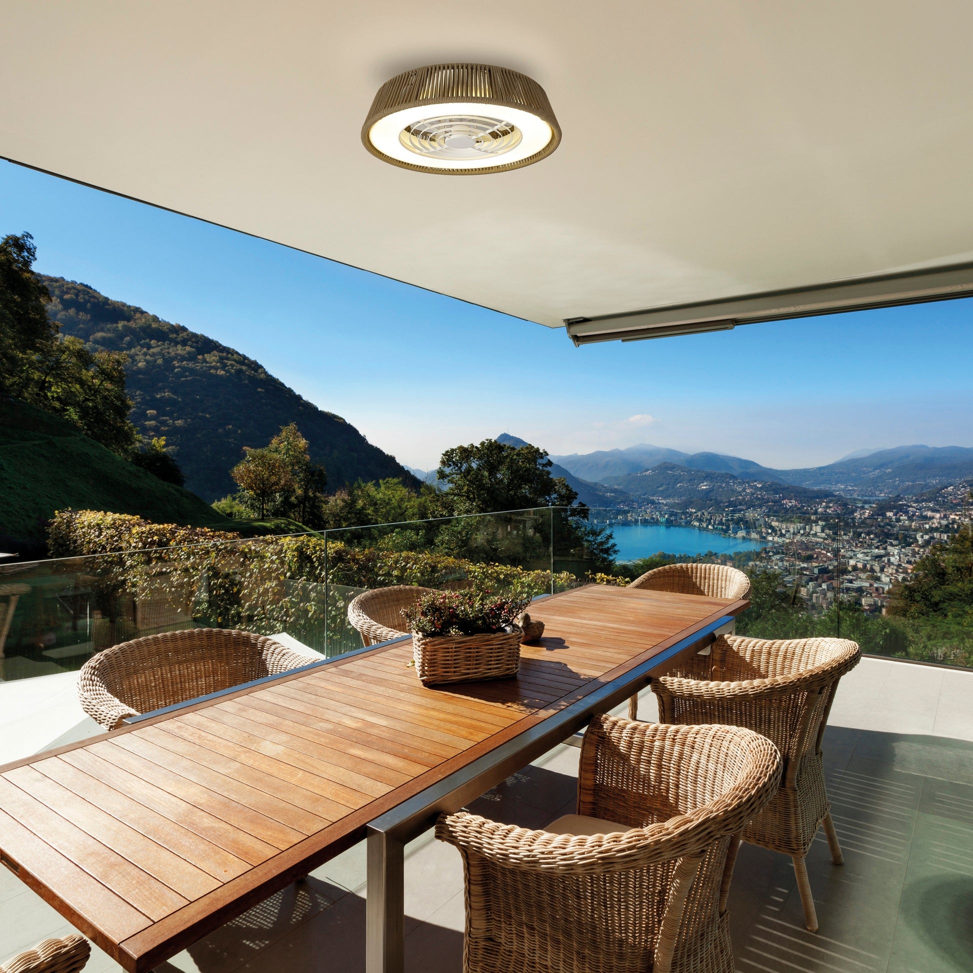 A spacious outdoor dining area features a wooden table surrounded by wicker chairs, set on a balcony with a glass railing. The balcony, enhanced by the Polinesia Nautica Mini 57.5cm 55W LED Dimmable Ceiling Light with Built-In 25W DC Reversible Fan from Ceiling Fans UK, offers a stunning view of lush green hills and a serene lake with a town nestled by its shores under a clear blue sky.