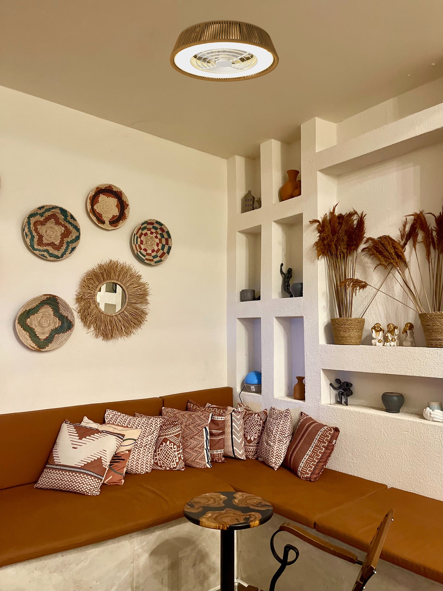A cozy corner with a brown cushioned seating area adorned with patterned pillows features decorative woven baskets hanging on the wall and built-in shelves displaying various ornaments and dried plants. Compact lighting, provided by the Polinesia Nautica Mini 57.5cm 55W LED Dimmable Ceiling Light With Built-In 25W DC Reversible Fan from Ceiling Fans UK, highlights the small wooden table and chair in front of the seating area.