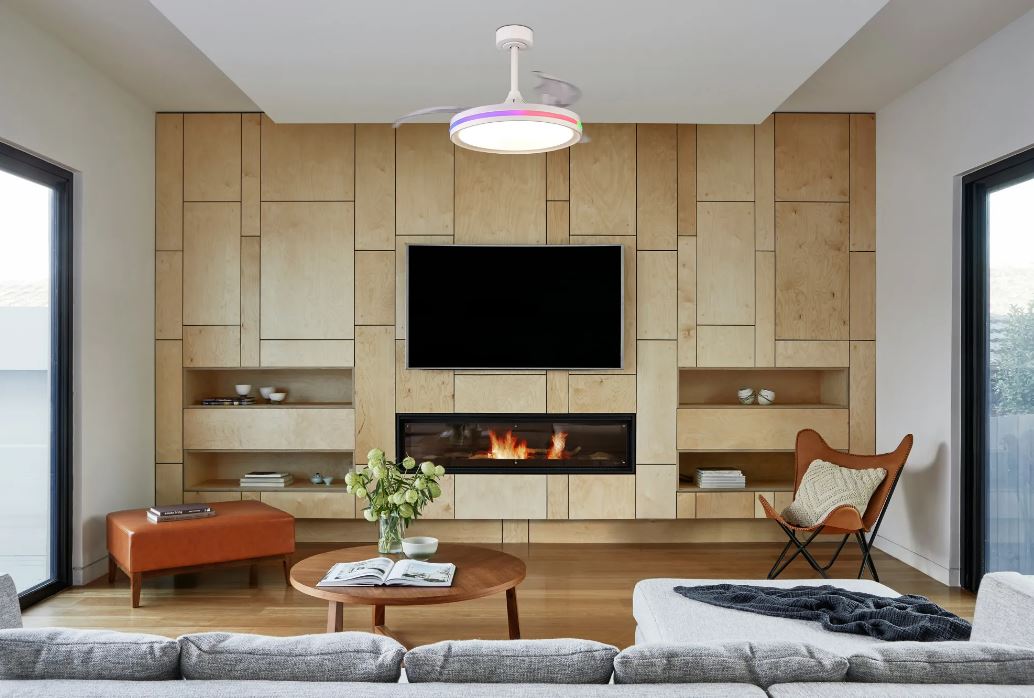 A modern living room with a minimalist design. The room features a wooden panel wall with a built-in fireplace and TV. A gray sofa and round wooden coffee table with a flower vase sit in the foreground, and a leather chair is in the corner. Large windows bring in natural light, complemented by the Rafaga 50W LED Dimmable Black/RGB Ceiling Light With Built-In 30W DC Reversible Fan from Ceiling Fans UK, offering adjustable brightness from 3000-6500K at 3200lm via remote control.