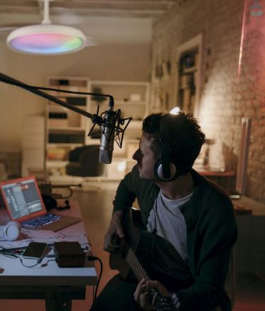 In a cozy room, a person wearing headphones strums an acoustic guitar with a microphone positioned in front of them. On the table, a laptop displaying music software is illuminated by the warm ambiance of the Siberia Black Ceiling Fan with Light from Ceiling Fans UK. This versatile fan features a 50W LED dimmable light with RGB capabilities and offers color temperatures ranging from 3000-6500K, enhanced by remote control functionality to perfectly set the mood.