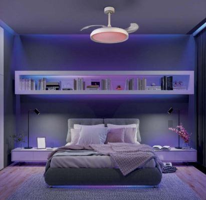 A modern bedroom with ambient lighting features a double bed, pillows, and a blanket. Hanging above is the Siberia Mini White Ceiling Fan with Light by Ceiling Fans UK, equipped with a 40W LED dimmable white/RGB ceiling light and 28W DC fan. The lighting offers an adjustable range between 3000-6500K and provides up to 2500lm brightness, all conveniently controlled by remote. Shelves filled with books line the wall, while two nightstands equipped with lamps are positioned on either side of the bed.