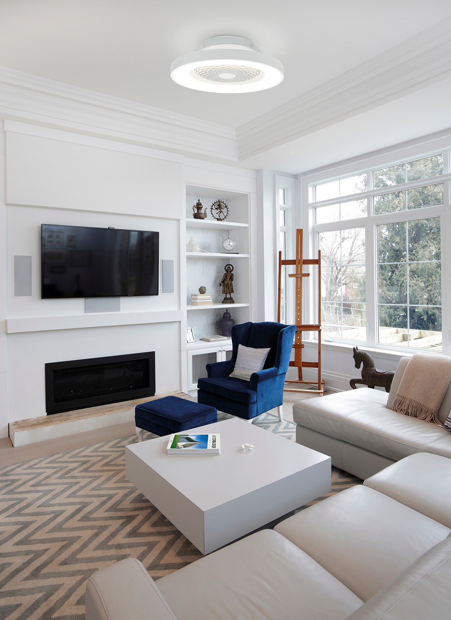 A modern living room features a white sectional sofa, a blue armchair with an ottoman, and a flat-screen TV mounted on the wall above a fireplace. Shelves with decorative items flank the TV. A Tibet White Ceiling Fan with Light 65cm 70W LED from Ceiling Fans UK adds ambiance, while a large window with trees outside provides natural light.