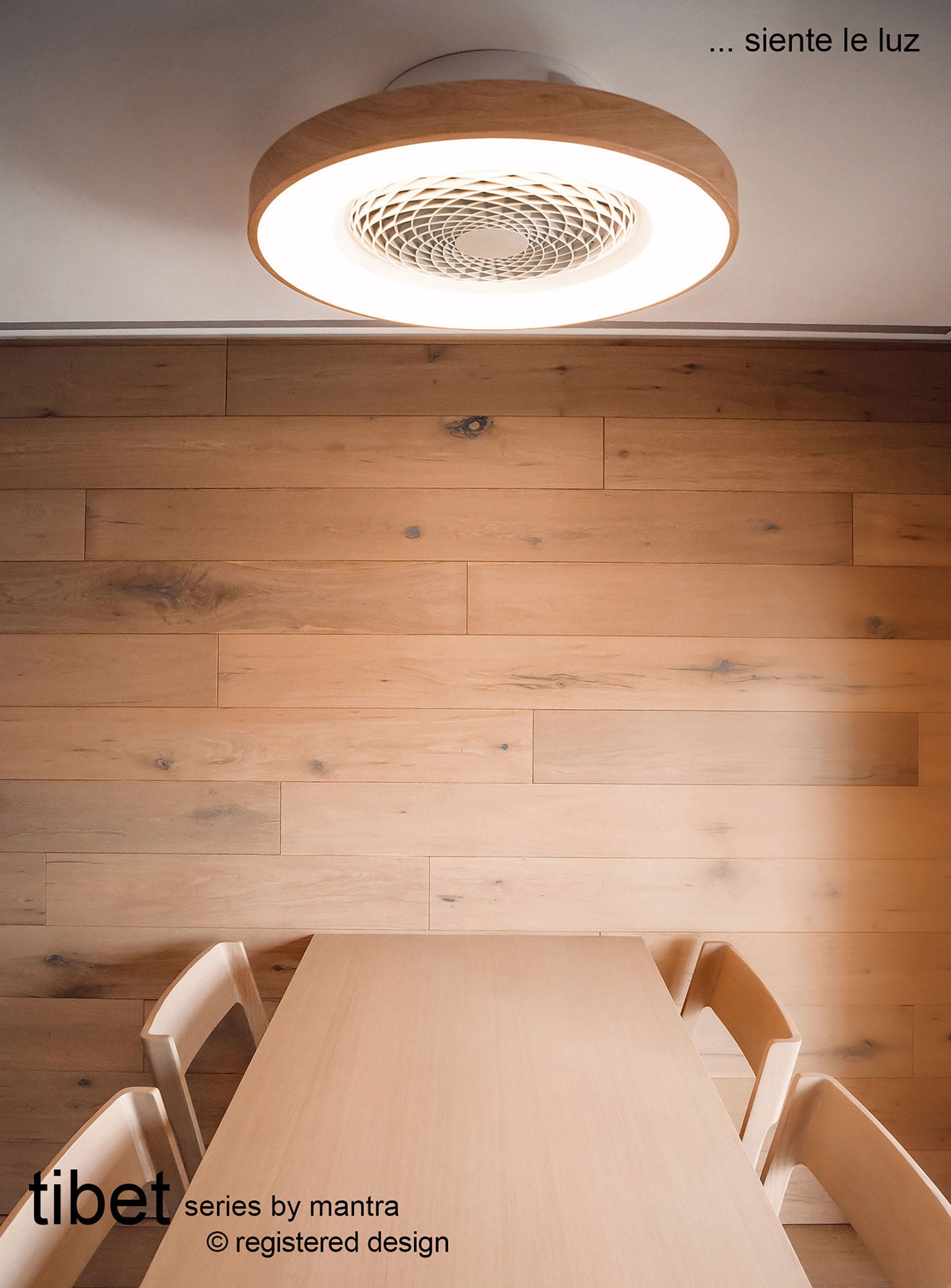 A minimalistic dining area with a wooden table and two wooden chairs. Above the table, a Tibet White/Wood Ceiling Fan with Light is mounted on the ceiling, featuring a 70W LED dimmable light that casts a warm glow. The fan, which includes a 35W DC reversible function, can be controlled via remote, app, or Alexa/Google Voice. The background features a wooden-paneled wall. Text reads: "Tibet series by Mantra © registered design" and "Siente le luz." Brought to you by Ceiling Fans UK with a 5-year warranty.