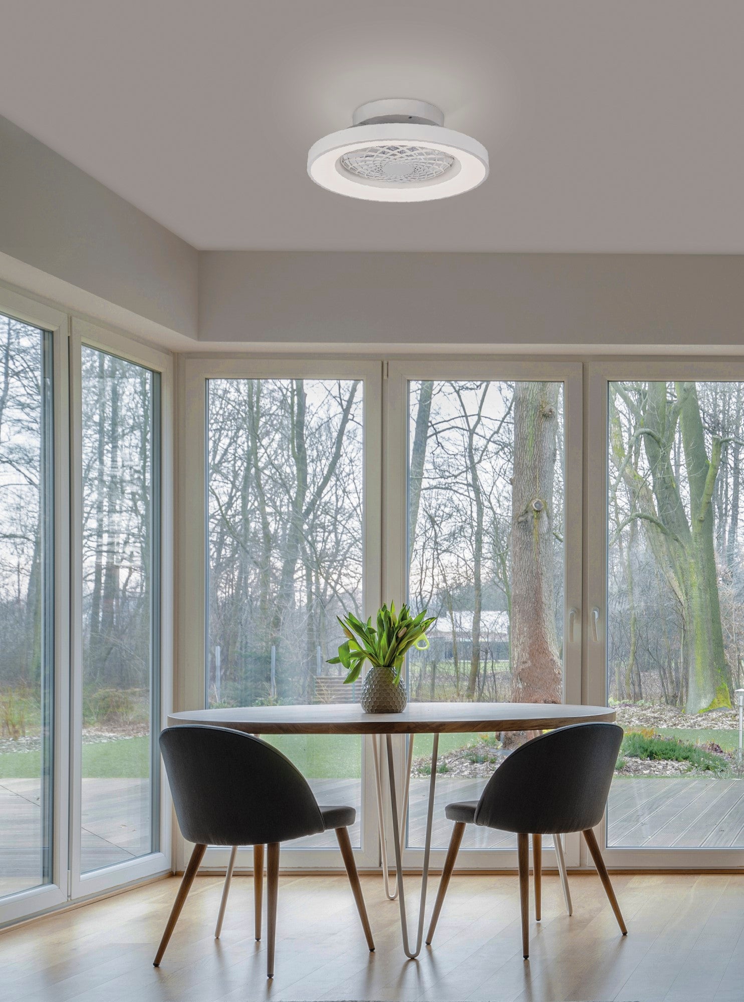 A dining area featuring a wooden round table with a vase of yellow flowers, flanked by two grey chairs. Tall windows and glass doors overlook a leafy backyard. A modern Ceiling Fans UK Tibet Mini White Ceiling Fan with Light (53cm, 70W LED Dimmable Ceiling Light with 35W DC Reversible Fan, Remote, 5yrs Warranty) illuminates the room perfectly.