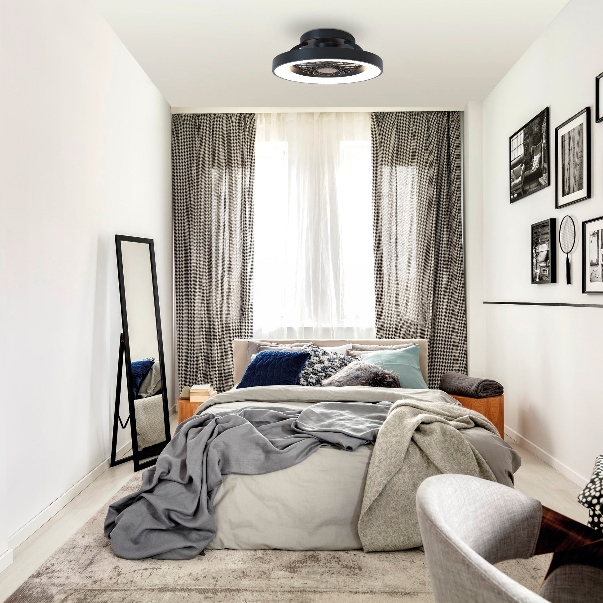 A cozy, minimalist bedroom features a bed covered in soft, gray bedding. A standing mirror is against the left wall, and framed pictures adorn the right. Natural light filters through sheer white curtains, while a Tibet Mini Black 53cm 70W LED Dimmable Ceiling Light With 35W DC Reversible Fan by Ceiling Fans UK completes the room's serene atmosphere.