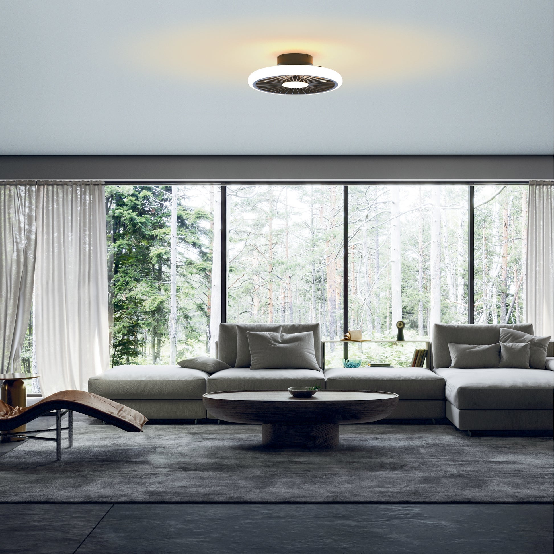 A modern living room with large floor-to-ceiling windows showcasing a forest view. The room features a beige sectional sofa with cushions, a round wooden coffee table, a lounge chair on the left, sheer white curtains, and a Turbo Black Ceiling Fan with Light by Ceiling Fans UK for perfect ambiance. This 51.2cm ceiling fan includes a 55W LED dimmable light and a built-in 30W DC reversible fan, backed by an impressive 5-year warranty.